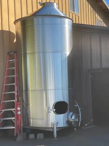 Stainless wine tank beside a winery building with a new oval port cut and fitted near the bottom, a ladder beside it